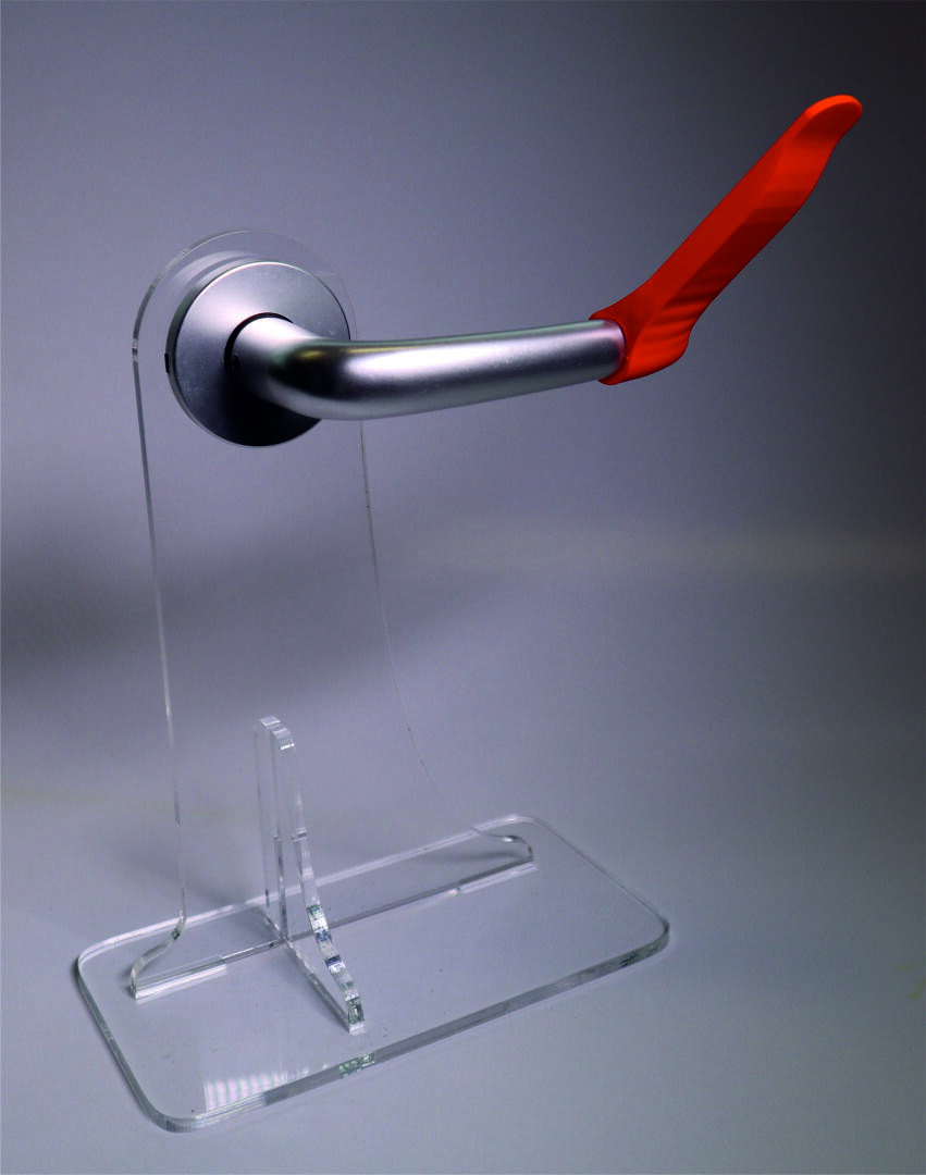 Ein Metalltürgriff mit einem roten Kunststoffaufsatz ist auf einem klaren Acrylständer vor einem neutralen Hintergrund angebracht.