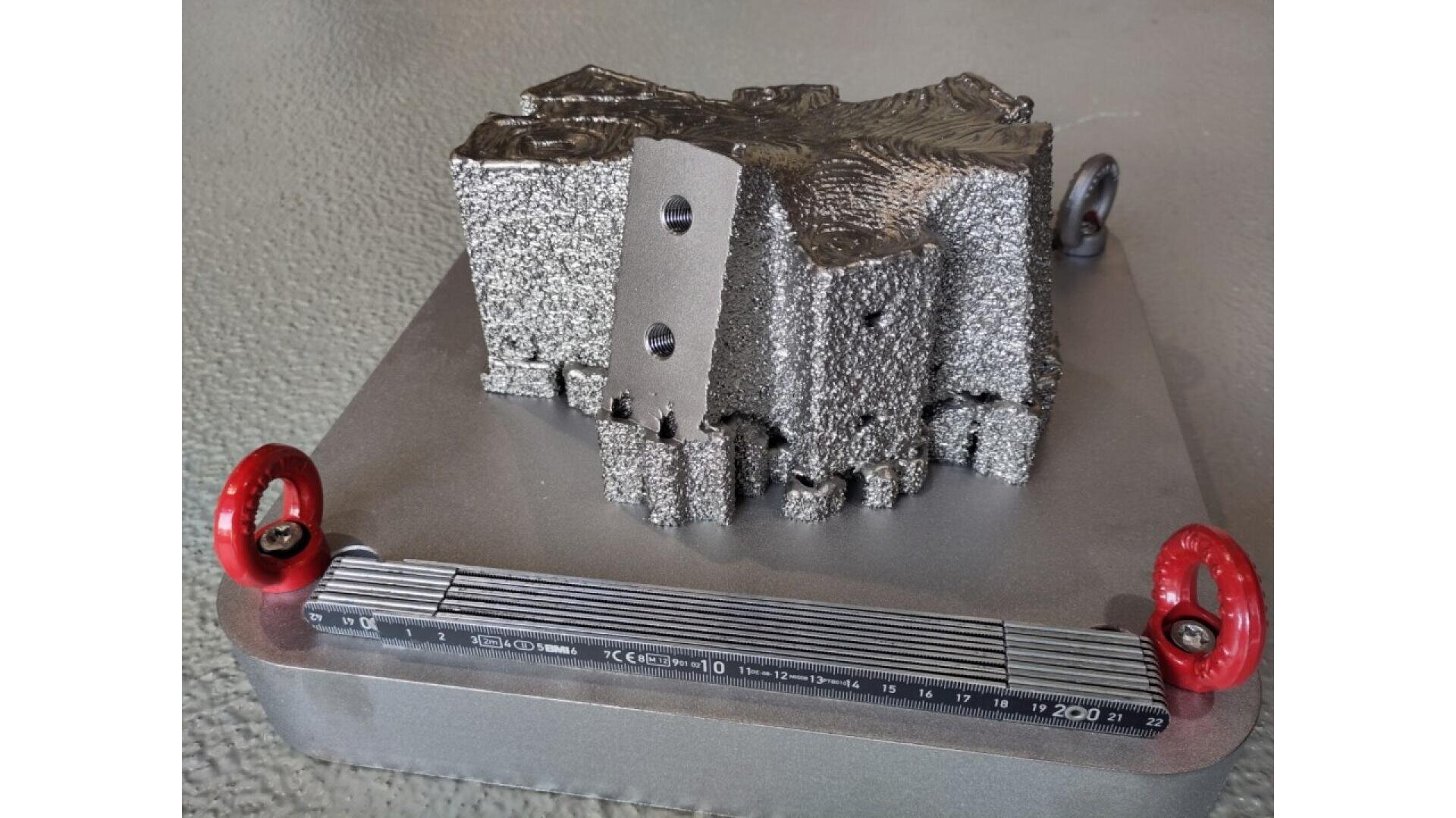 A complex, metallic 3D-printed object with rough and smooth surfaces is mounted on a square metal base with red handles and a ruler along the front edge. The background is a textured gray surface.