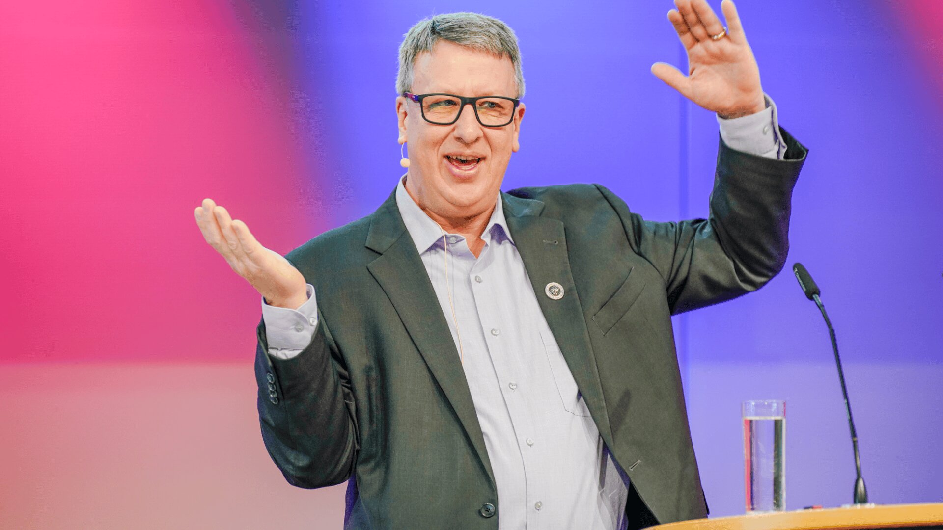 Ein Mann in Anzug und Brille gestikuliert enthusiastisch, während er an einem Podium mit einem Mikrofon und einem Glas Wasser vor einem bunten, unscharfen Hintergrund spricht.