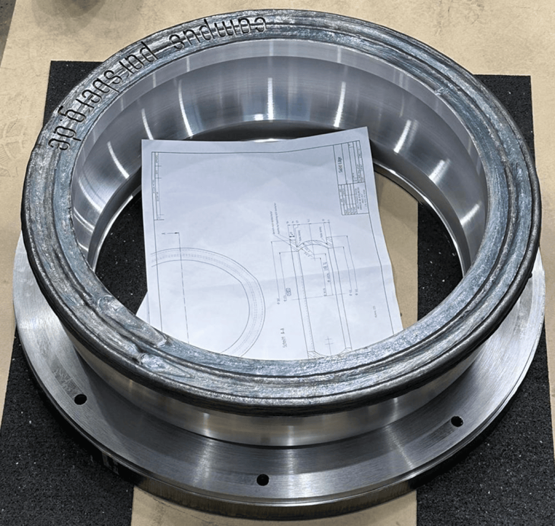 Ein blaugrau beschichteter Metallring liegt aufrecht auf einem Tisch und verdeckt teilweise eine technische Zeichnung mit technischen Maßangaben. Zum Schutz liegt der Ring auf einer dunklen Schaumstoffunterlage.