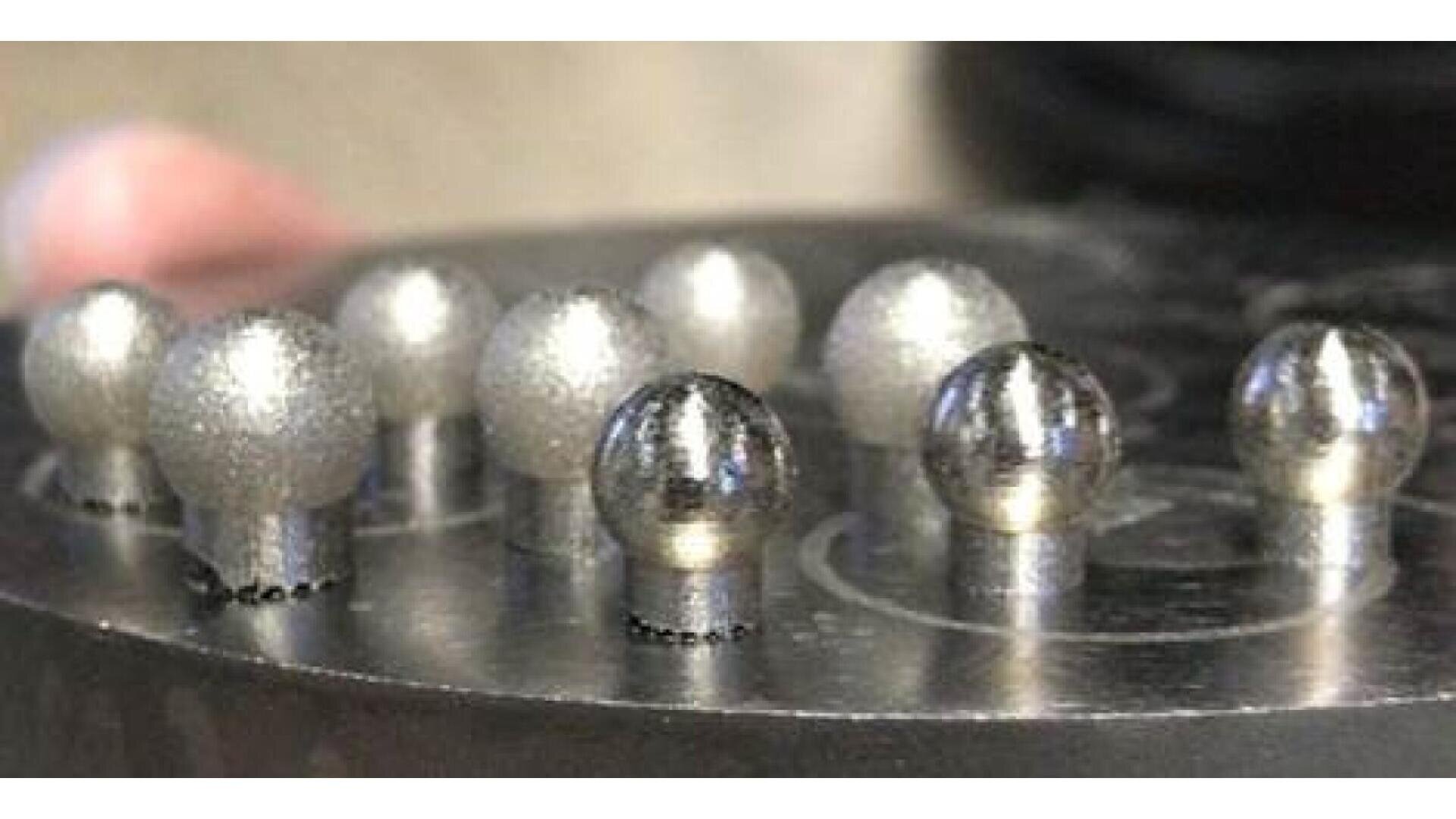 A close-up of nine metallic ball-shaped objects arranged in rows on a circular metal surface. Some balls have a rough, frosted texture while others appear smooth and shiny.