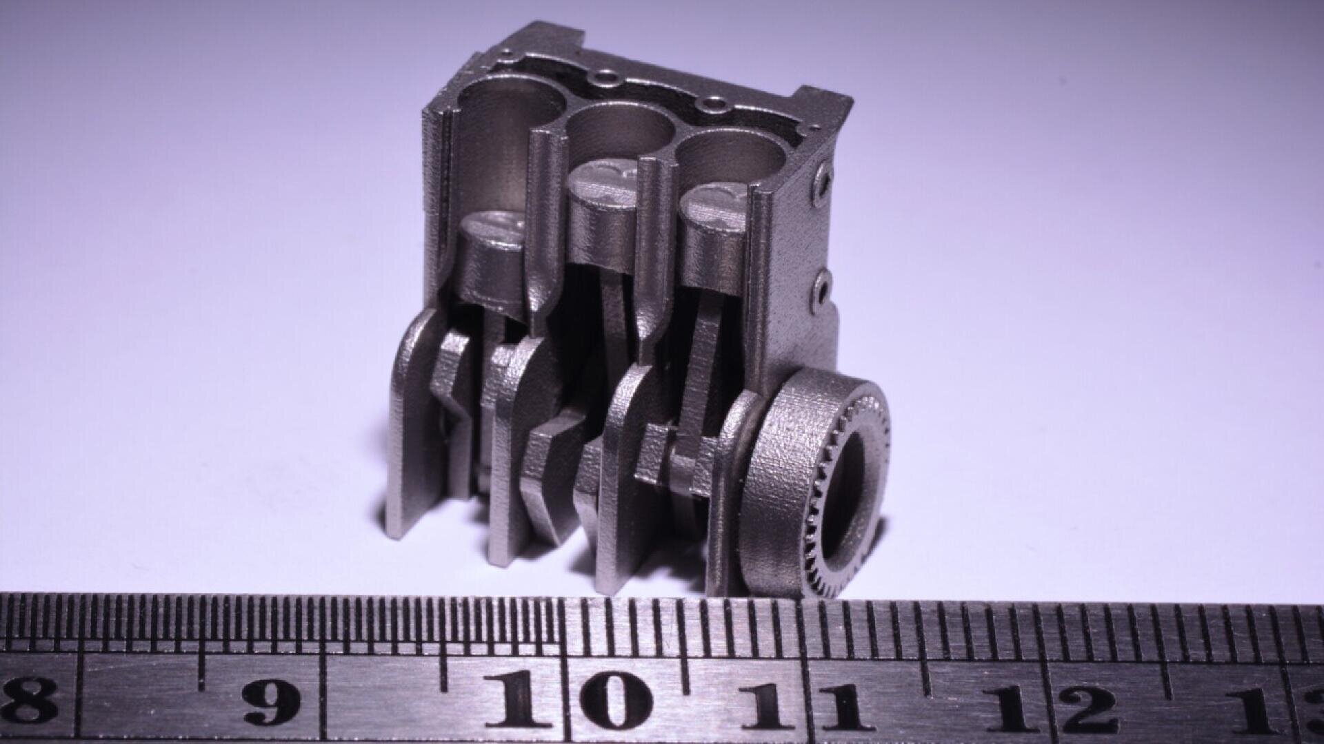 A small, intricate metal mechanical part resembling an engine or gear component is placed on a white surface next to a ruler, showing it is about 3 centimeters tall.