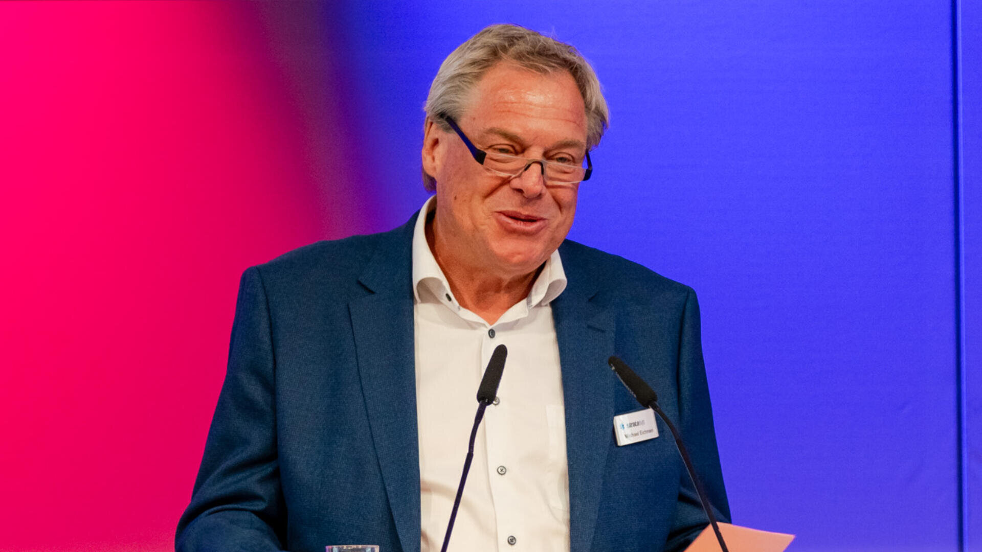 Ein älterer Mann in blauem Anzug und weißem Hemd spricht lächelnd an einem Podium mit zwei Mikrofonen. Der Hintergrund ist in Rot und Blau gehalten. Er trägt eine Brille und ein Namensschild.