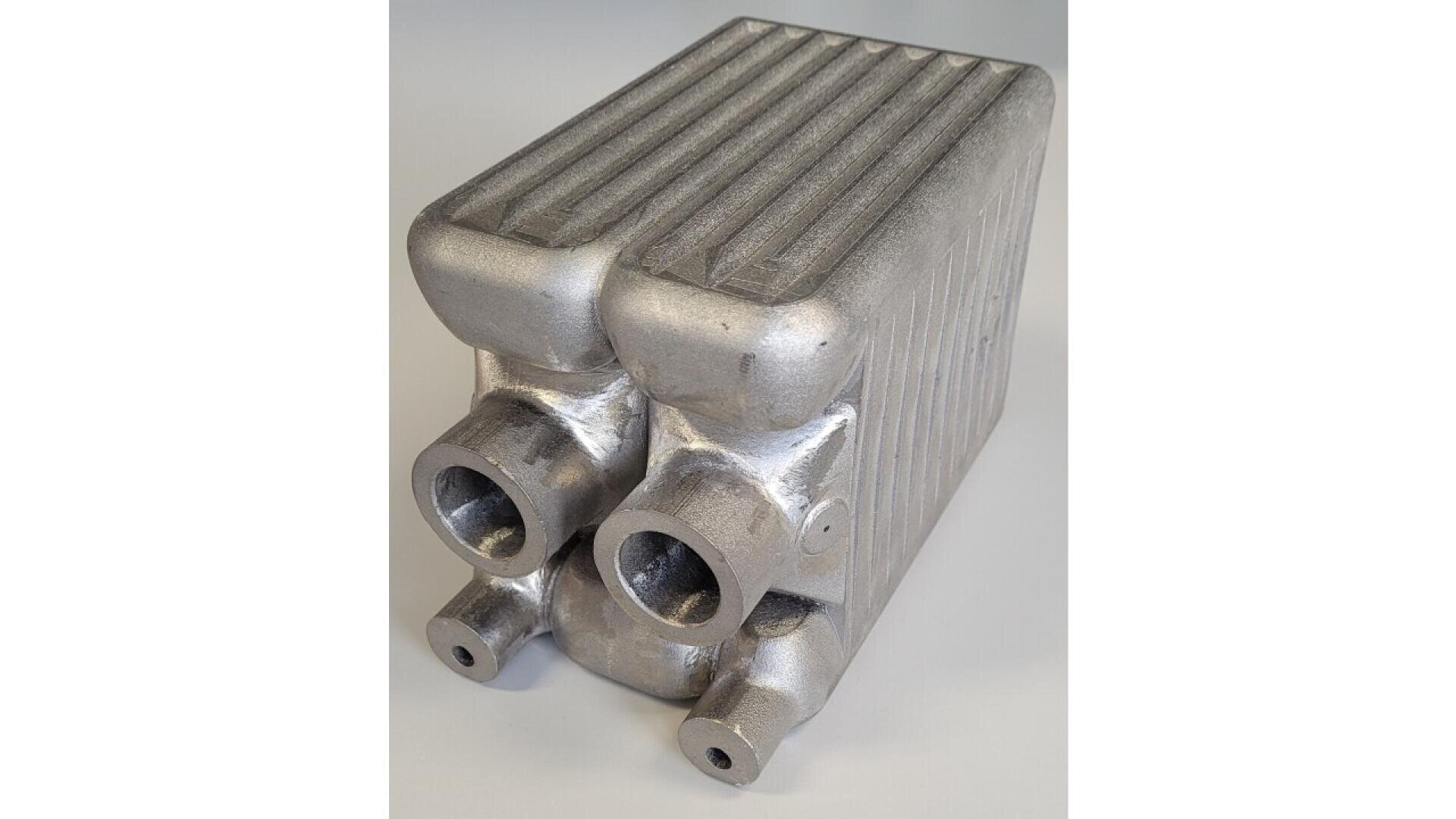 A metallic, rectangular heat exchanger with ridged top surface and four cylindrical pipe inlets or outlets on one end, designed for industrial fluid or gas transfer.