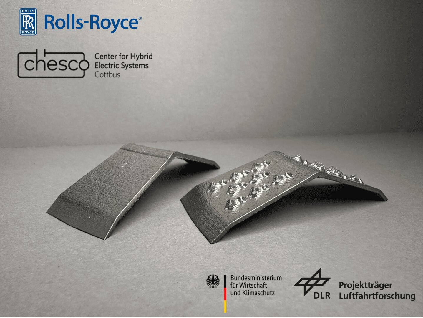 Auf einer grauen Fläche liegen zwei Bleche; eines ist glatt, das andere weist mehrere Erhebungen auf. Ober- und unterhalb der Bleche sind die Logos von Rolls-Royce, Chesco, dem Bundesministerium für Verkehr und digitale Infrastruktur (BMI) sowie der DLR zu sehen.