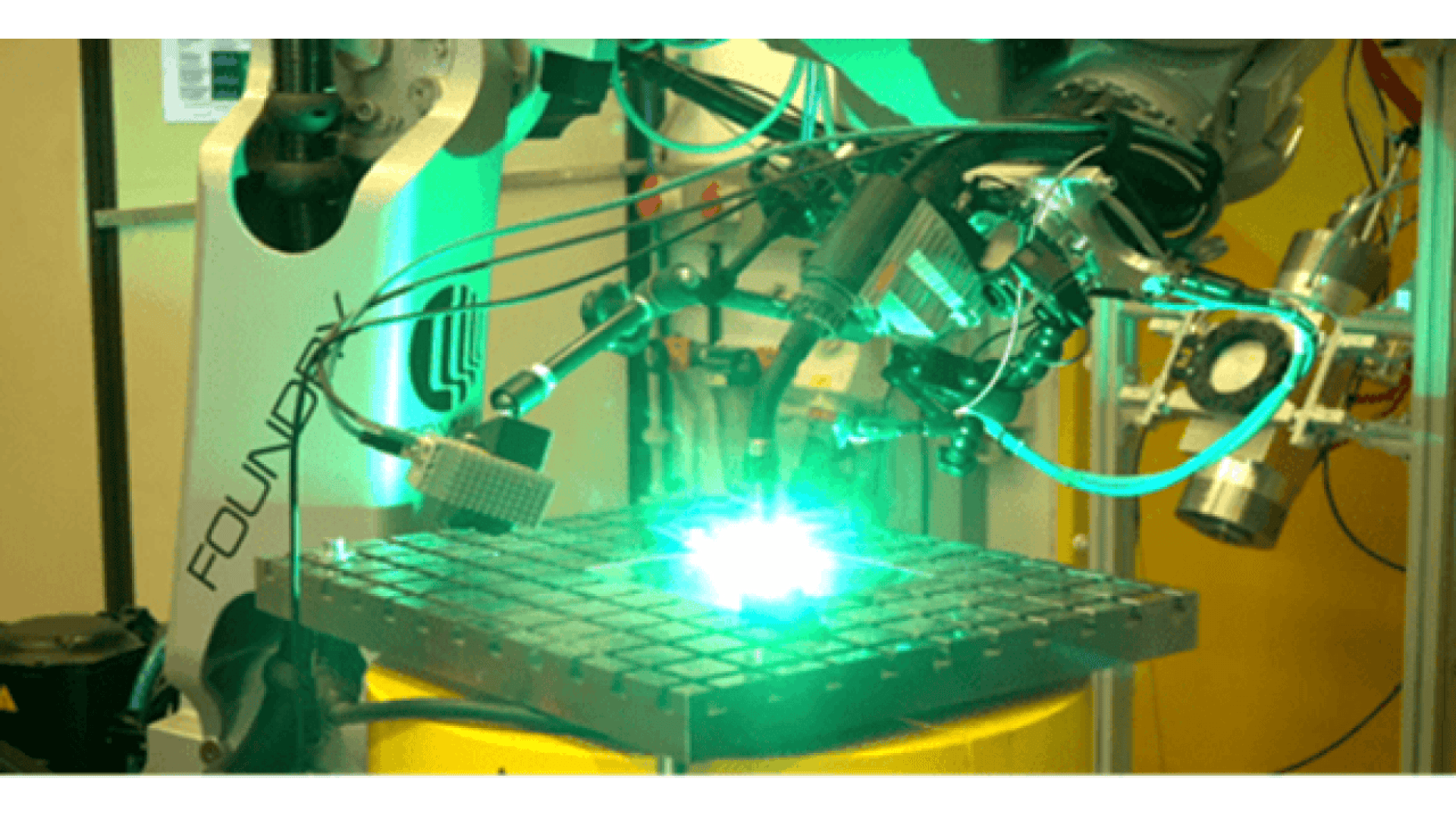 A robotic arm equipped with tools and sensors performs a bright, illuminated task on a metal grid in a laboratory setting with yellow and green lighting.