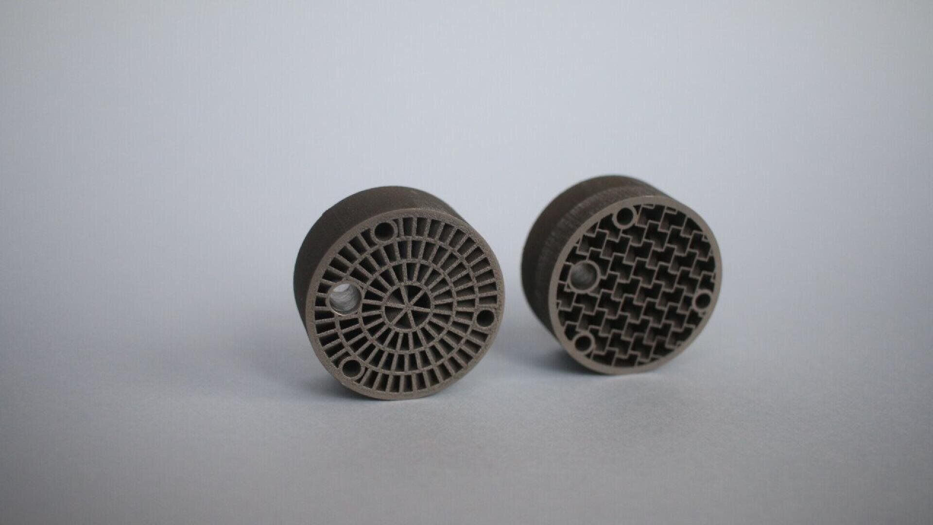 Two round, dark gray 3D-printed objects are shown on a light gray surface. Each has a different geometric pattern of holes on the front face—one with a radial design, the other with a grid-like structure.
