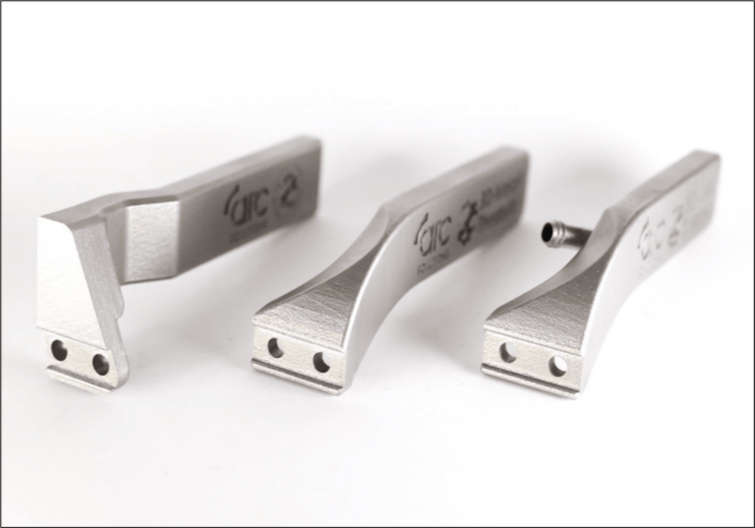 Three metallic, precision-machined components with engraved markings are shown standing on a white surface. Each piece has a flat base with two small holes and a uniquely shaped, angled body.