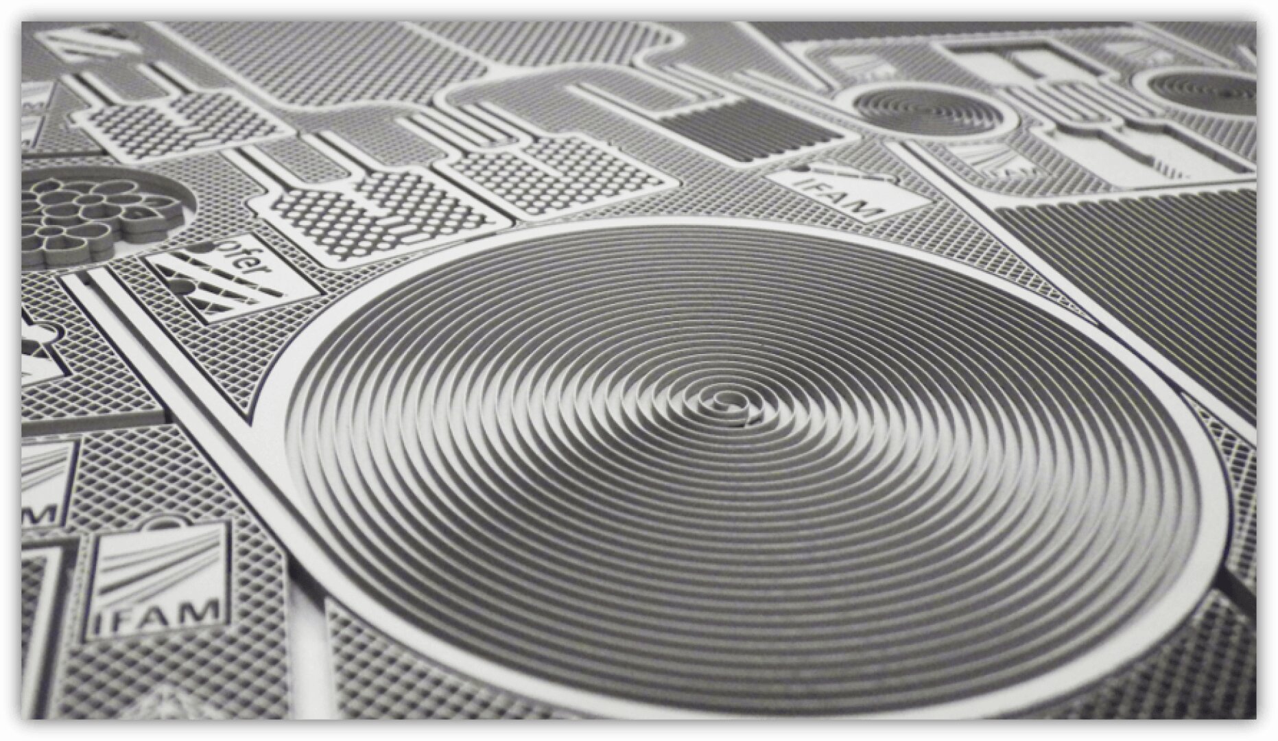 Nahaufnahme einer metallischen Leiterplatte mit verschlungenen Mustern, mit einem großen kreisförmigen Spiralmuster in der Mitte und verschiedenen strukturierten Komponenten drum herum. Das Bild ist monochromatisch und sehr detailliert.