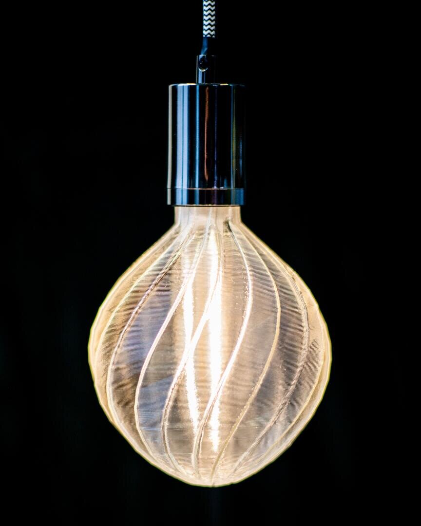 A glowing, modern light bulb with a swirling, textured glass design hangs against a solid black background. The filament is illuminated, casting a warm, soft light within the bulb.