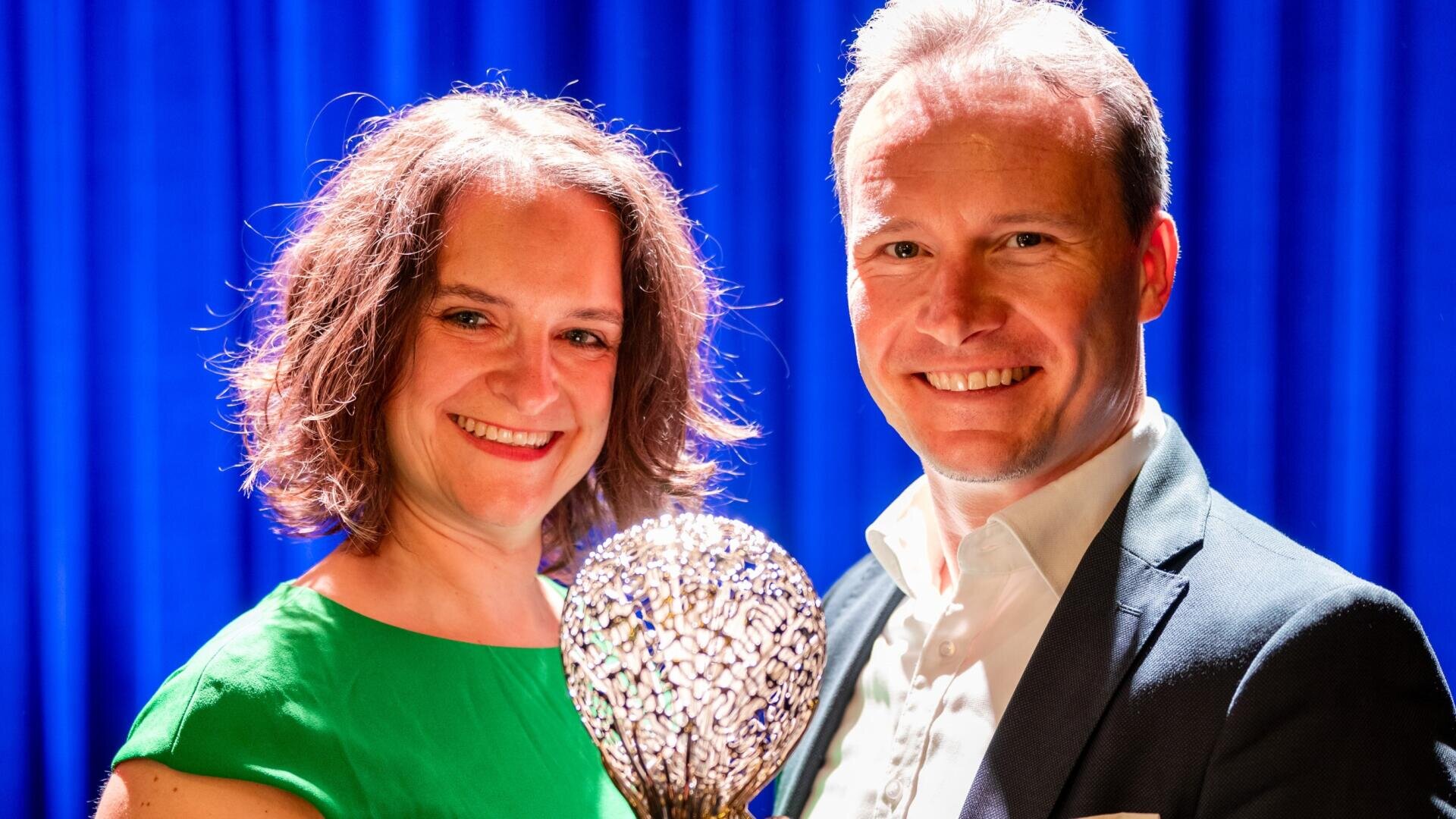 Eine Frau im grünen Kleid und ein Mann im Anzug lächeln, während sie eine dekorative Trophäe halten und vor einem blauen Vorhang mit Bühnenbeleuchtung stehen.