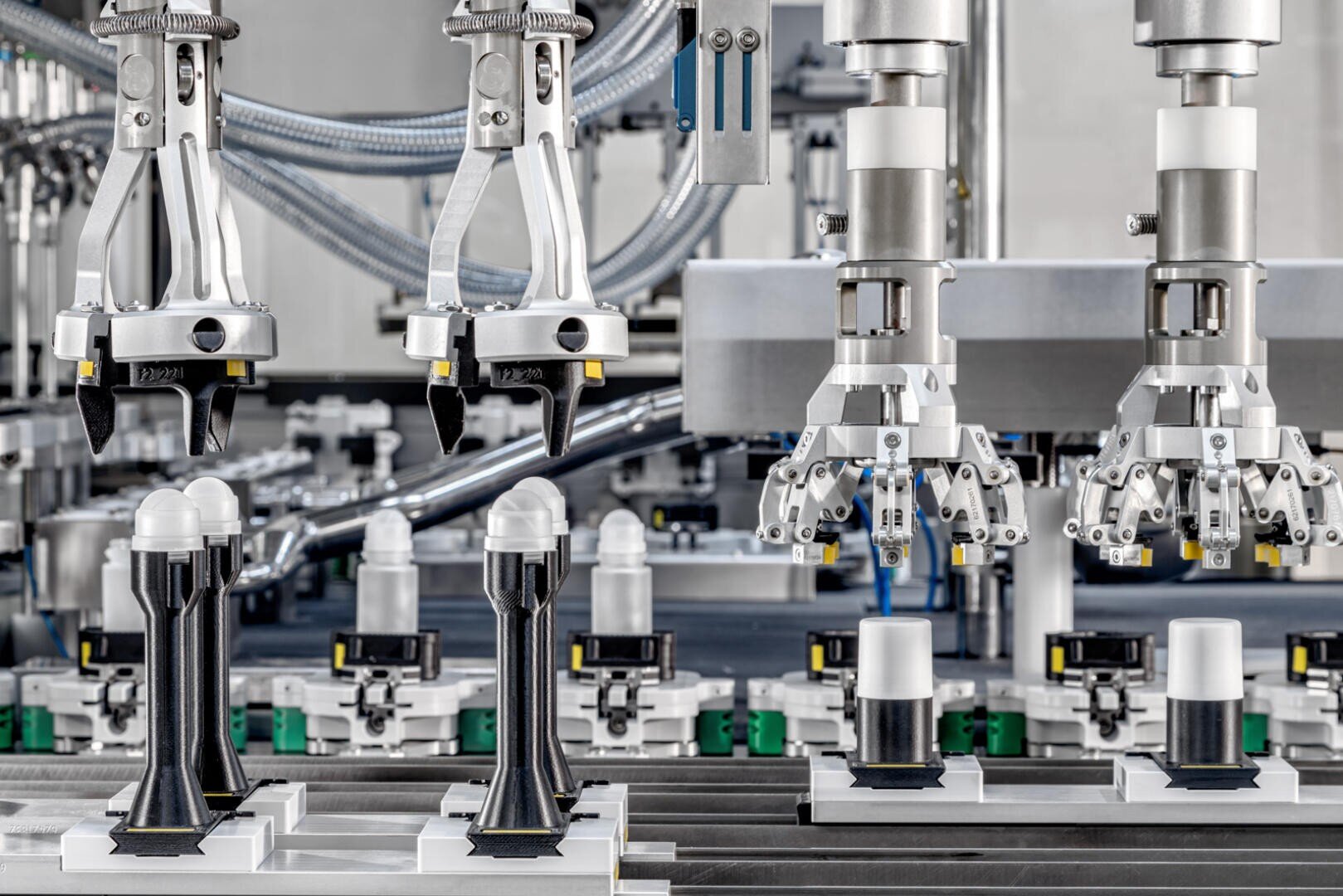 Roboterarme in einer Fabrikmontagelinie handhaben und platzieren zylindrische weiße Objekte präzise auf schwarzen Ständern, mit verschiedenen mechanischen Teilen und Förderbändern im Hintergrund.
