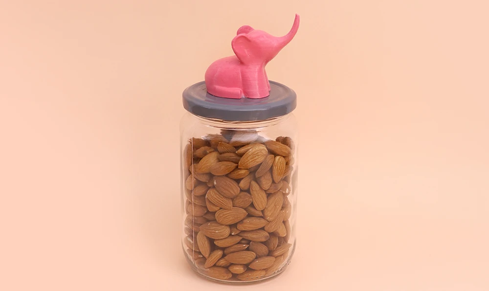 A glass container with almonds, the lid of which is decorated with a 3D-printed pink elephant figure.