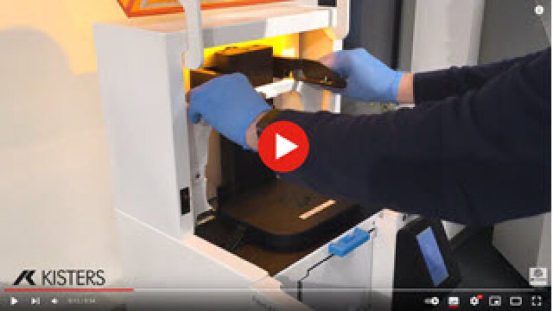 Eine Person mit blauen Handschuhen bedient einen 3D-Drucker mit einer orangefarbenen Abdeckung. Der Drucker ist teilweise geöffnet, und die Person scheint ein Bauteil einzulegen oder zu entfernen. Das Kisters-Logo ist in der unteren linken Ecke zu sehen.