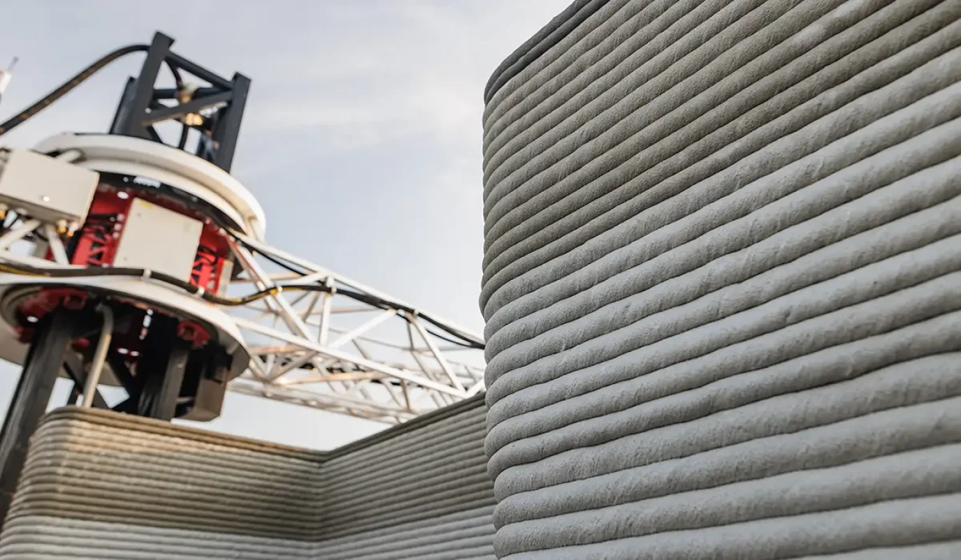 Ein großformatiger 3D-Drucker trägt schichtweise Beton auf und errichtet so die Wände eines Gebäudes – eine moderne Anwendung des 3D-Drucks im Bauwesen.