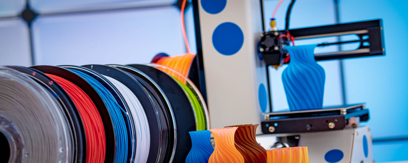 Several colorful filament spools stand side by side, while a 3D printer is forming a blue-printed vase next to them. Other colorful vases with curved designs lie on the table.