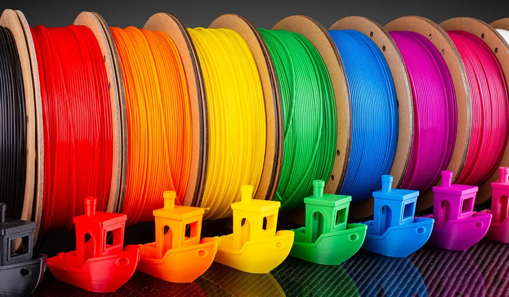 Bunte Filamentspulen in Regenbogenfarben stehen nebeneinander, davor sind passende kleine 3D-gedruckte Boote in denselben Farben aufgereiht. Die Szene wirkt geordnet und farbenfroh.