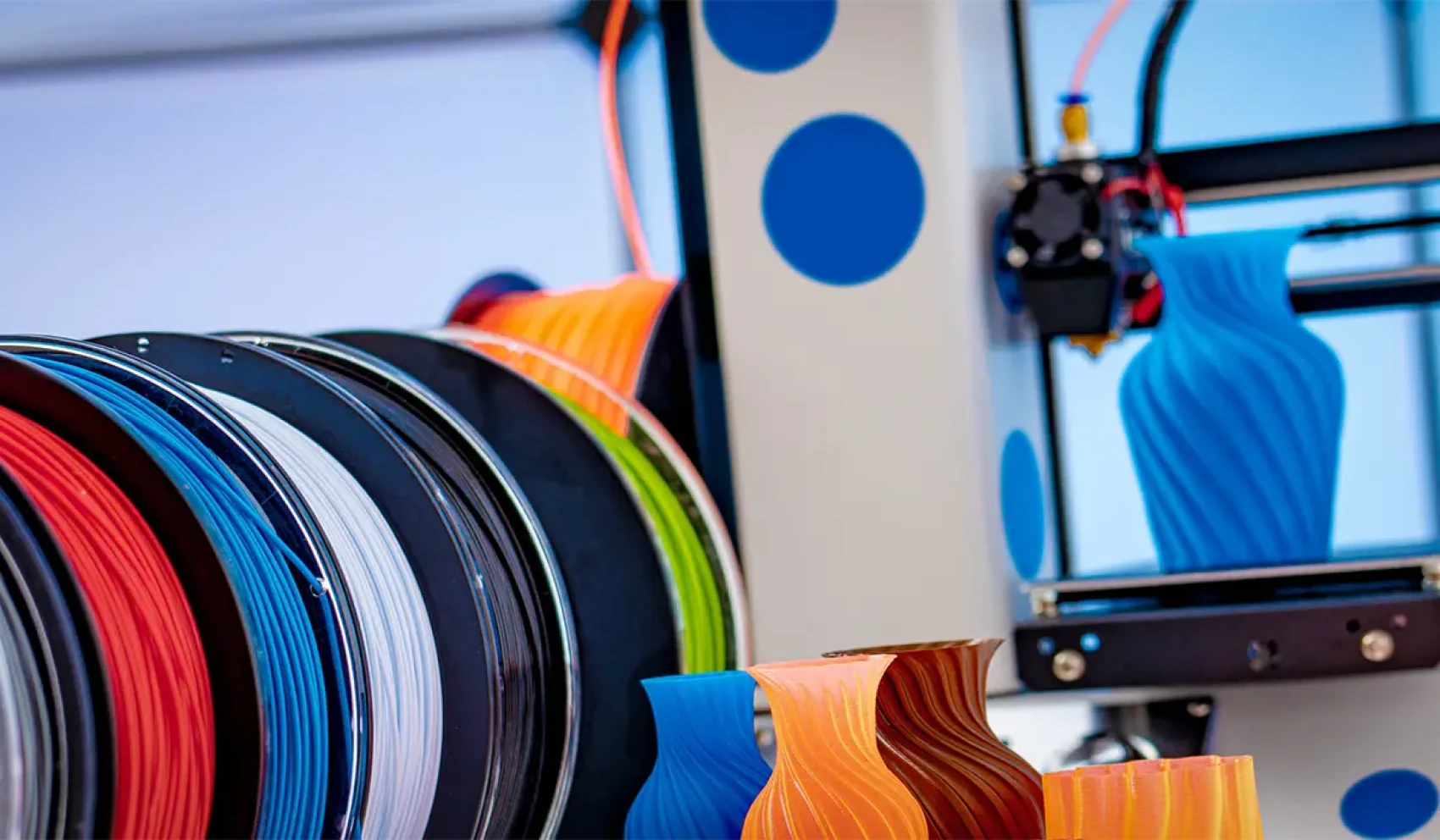 Mehrere bunte Filamentrollen stehen nebeneinander, während daneben ein 3D-Drucker eine blau gedruckte Vase formt. Auf dem Tisch liegen weitere farbige Vasen mit geschwungenem Design.