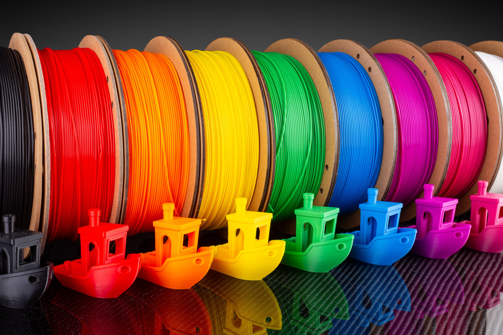 Colorful filament spools in rainbow colors stand side by side, with matching small 3D-printed boats in the same colors lined up in front of them. The scene appears orderly and colorful.